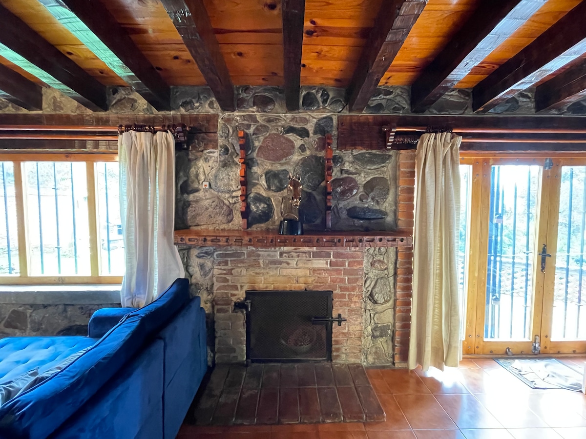 A cozy living area features a stone wall with a rustic fireplace, complemented by wooden beams overhead. Large windows invite natural light, while light curtains frame the view. A plush blue couch provides a relaxed seating option, enhancing the inviting atmosphere.