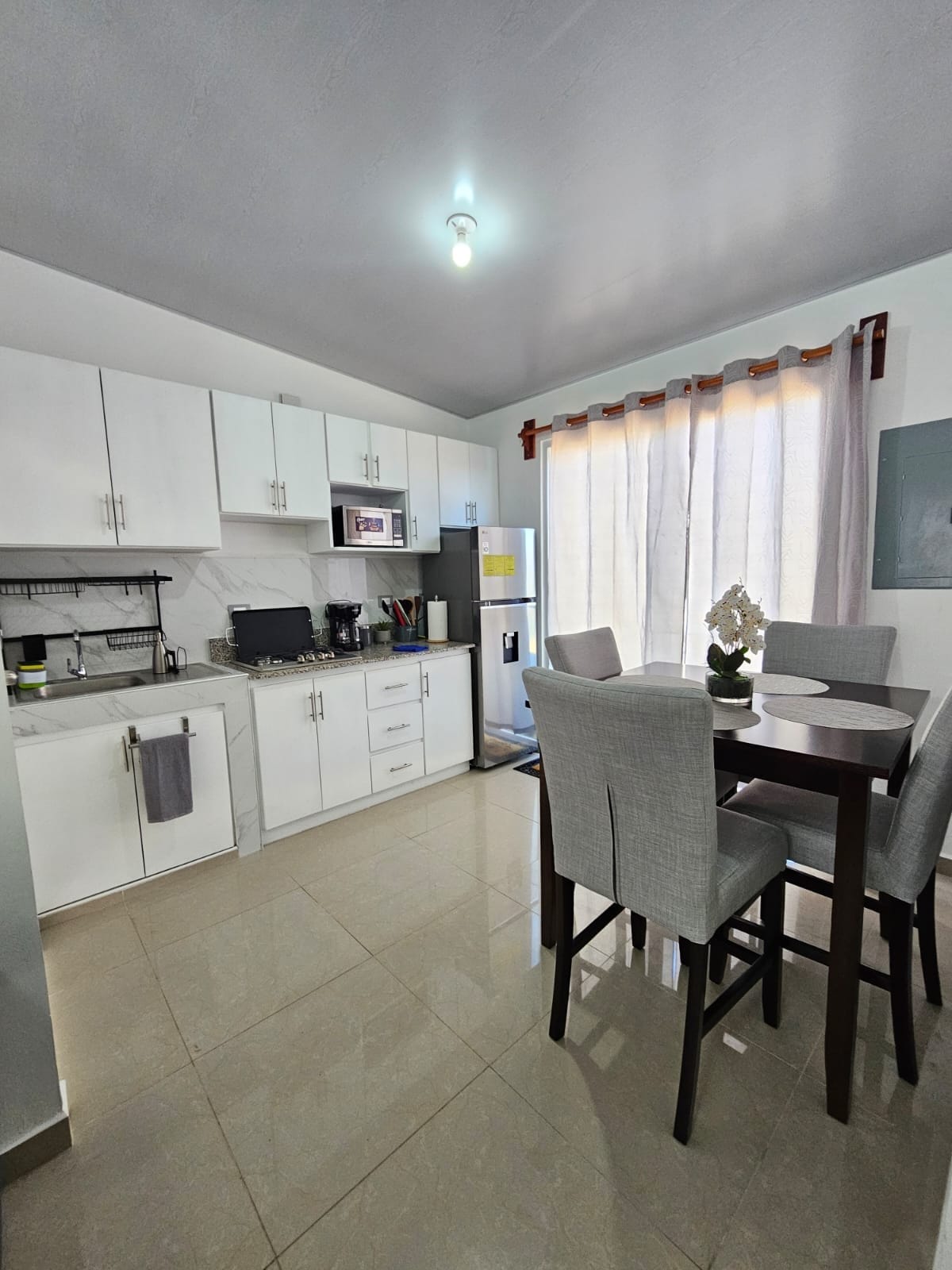 A modern kitchen is shown, featuring sleek white cabinetry and appliances. A dining table with four upholstered chairs sits near large windows, allowing natural light to brighten the space. The kitchen counter is equipped with essential appliances, enhancing functionality.