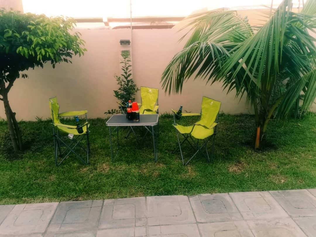 A small outdoor seating area is depicted, featuring four bright green folding chairs arranged around a simple table. Grassy ground is visible between the chairs, with two leafy palm trees in the background and a low, neutral-colored wall partially enclosing the space.
