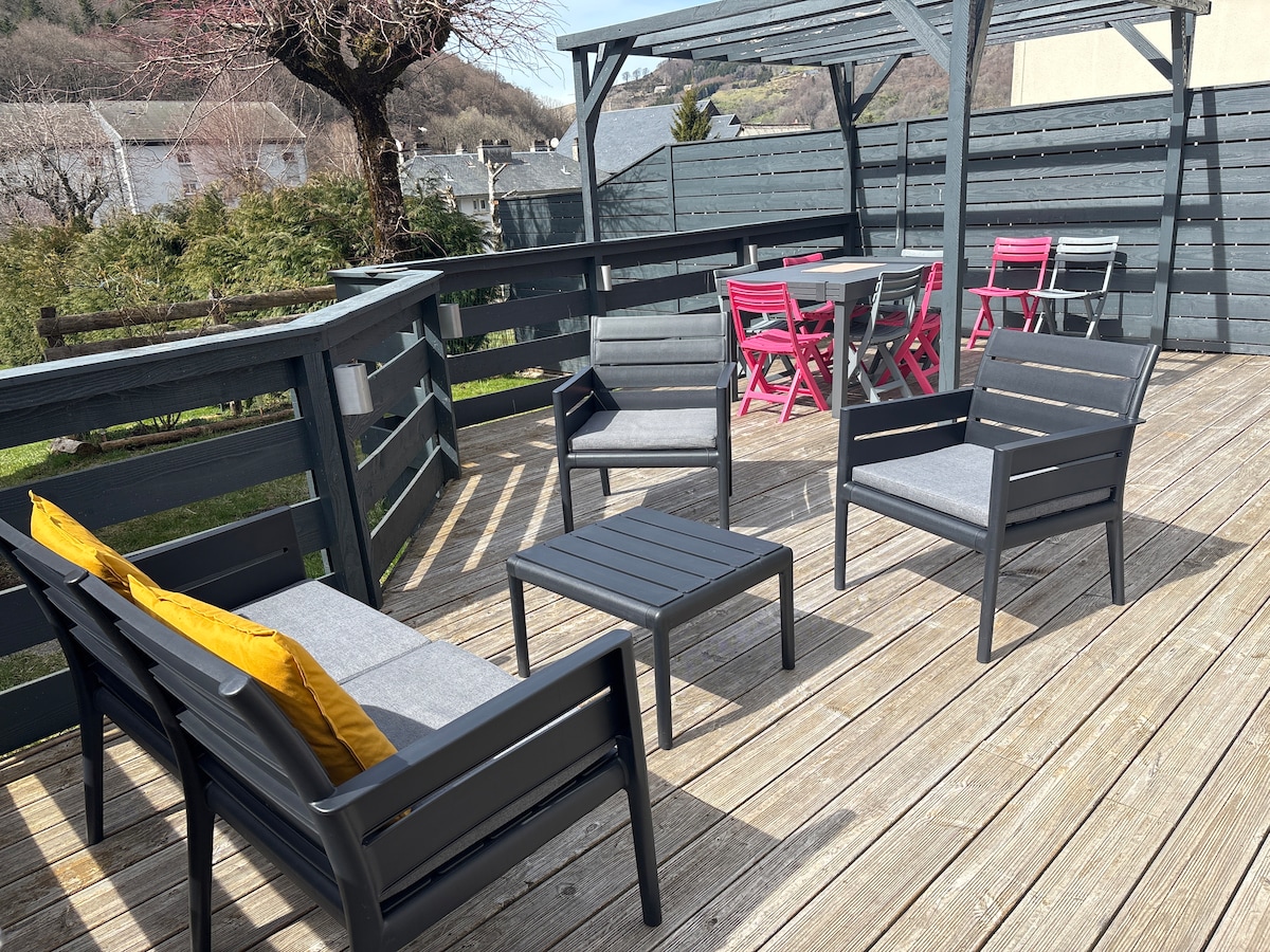A wooden terrace features several outdoor seating options, including two armchairs and a low table with a single yellow accent pillow. A dining space is visible with a table and chairs in shades of pink. The surrounding greenery offers a natural backdrop.