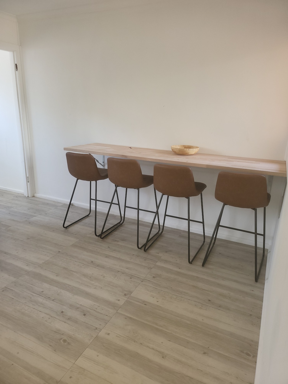 A sleek breakfast bar is positioned against a neutral wall, accompanied by four ergonomic, brown high chairs with slender black legs. The wood grain of the bar contrasts with the pale floor, contributing to a modern, functional dining area.