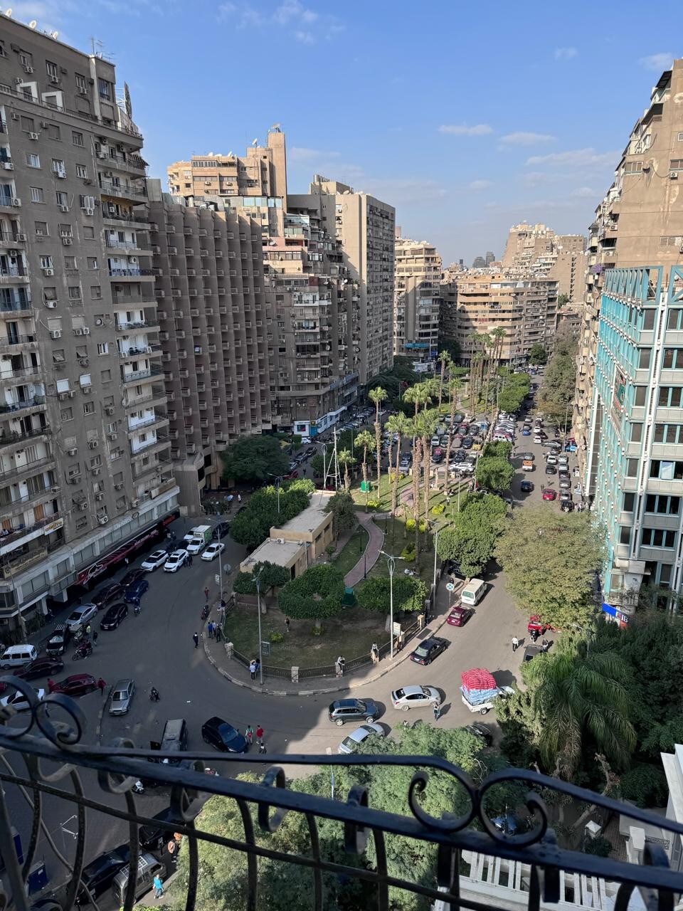 An aerial view captures the bustling cityscape, showcasing a vibrant green park at the center, surrounded by tall buildings. Cars are visible along the winding streets, while palm trees and open spaces add a touch of nature to the urban environment.