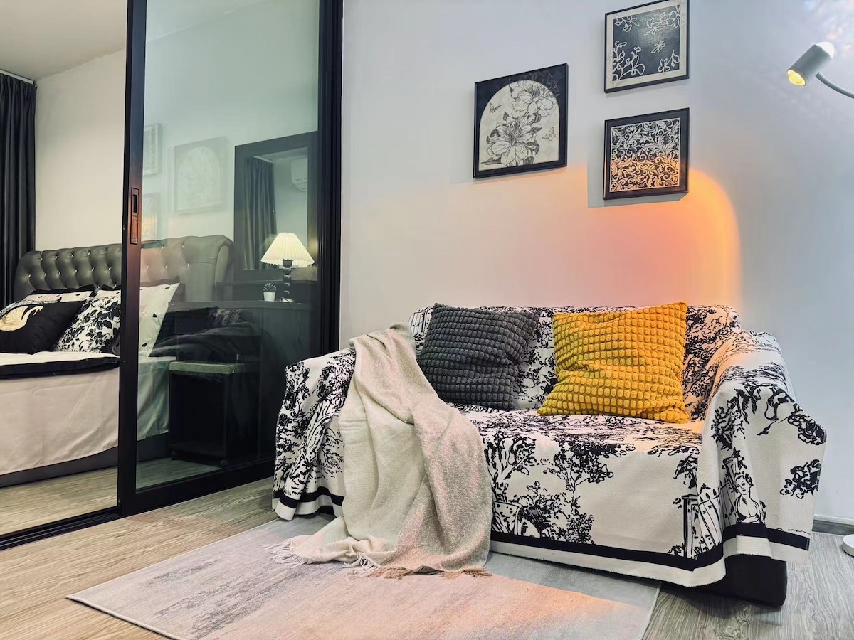 A comfortable seating area is presented with a patterned sofa draped with a soft throw. Decorative pillows add color, while framed art hangs on the wall. A glimpse of the bedroom can be seen through the glass partition, creating an open feel.