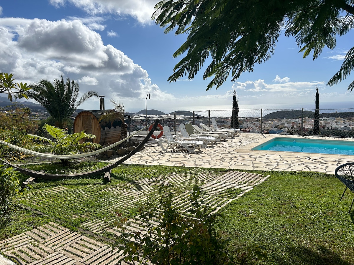 An outdoor space showcases a swimming pool surrounded by lounge chairs. A hammock is draped between two trees, offering a relaxing area. Lush greenery and terracotta pathways lead to a distant view of hills and clouds, enhancing the serene environment.