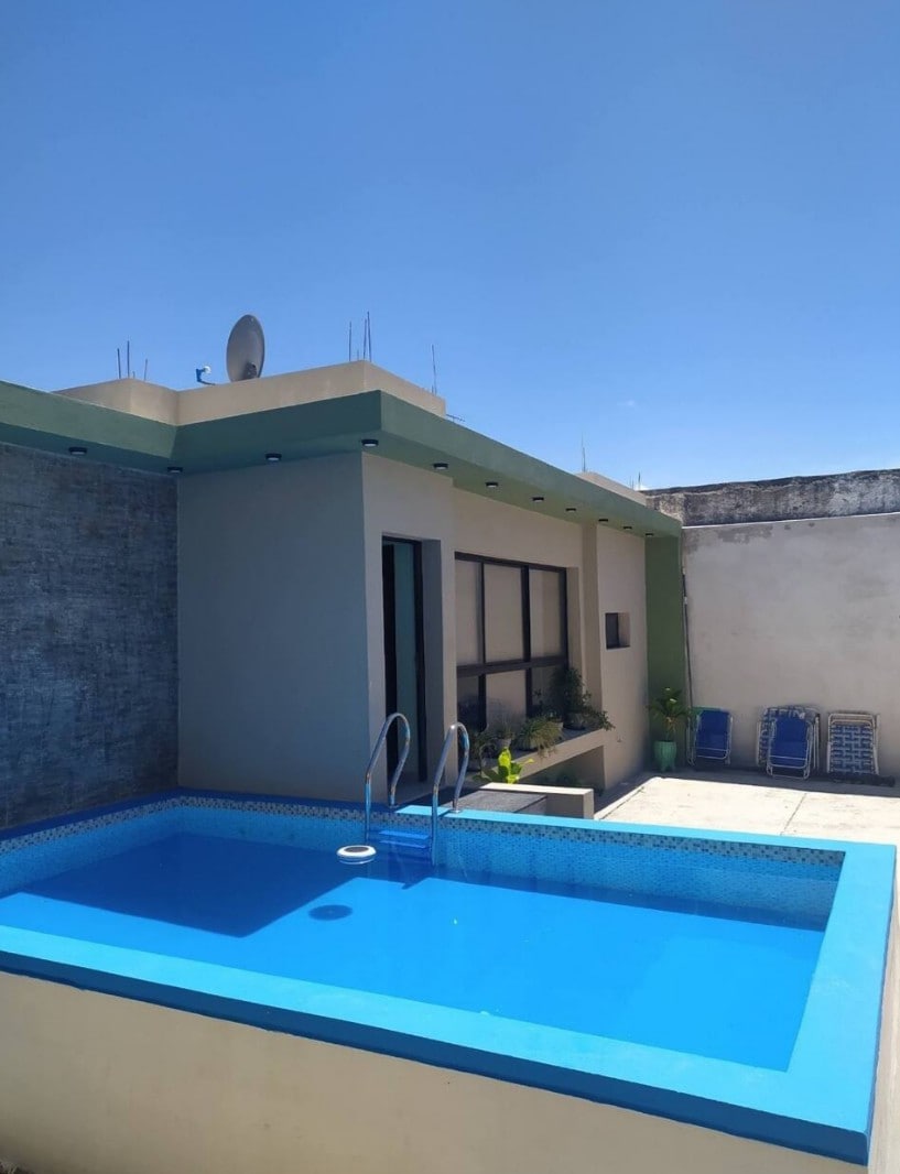 A bright outdoor space is showcased, featuring a small swimming pool surrounded by blue tiles. Lounge chairs are arranged nearby, and an exterior wall displays a textured finish. Clear skies are visible above, enhancing the openness of the area.