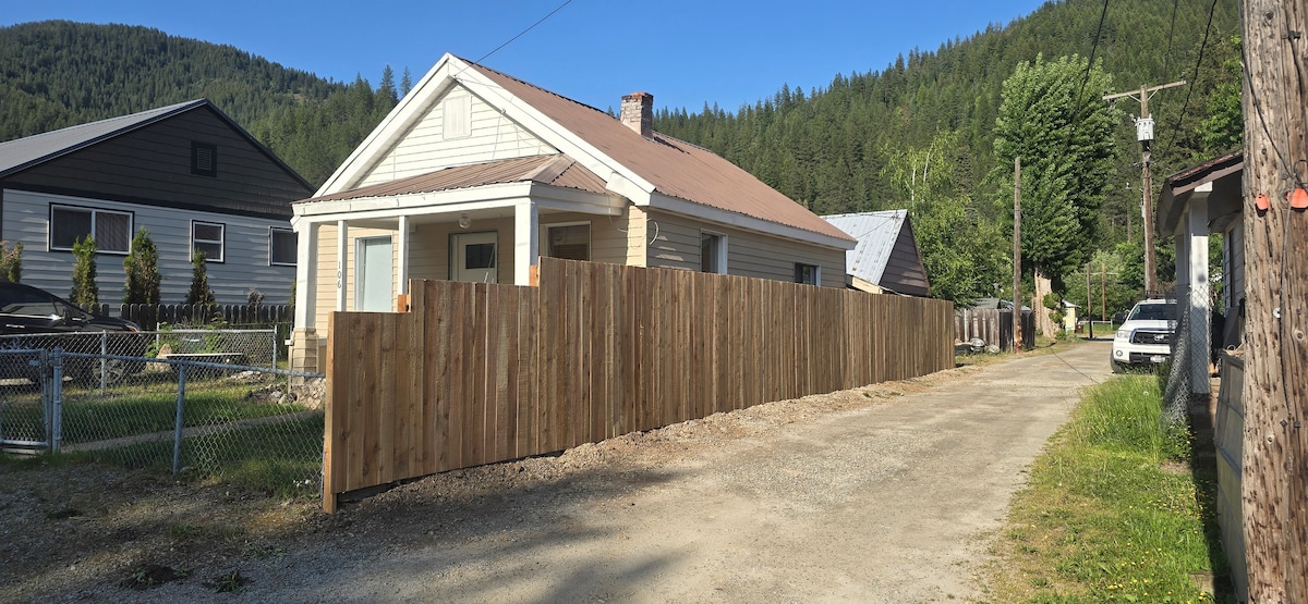 A single-story home with a light-colored exterior is framed by a wooden privacy fence. The property features a gravel driveway bordered by greenery, with mountains rising in the background under a clear blue sky. Nearby structures include additional residential homes and a view of a tree-lined area.