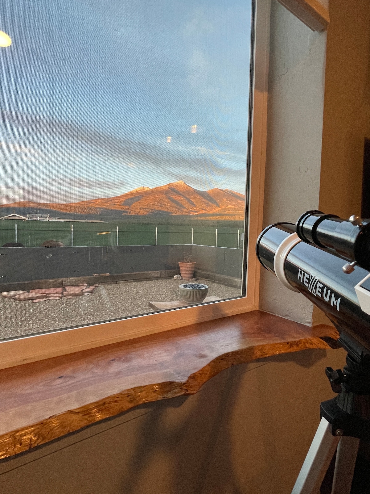 A telescope is positioned beside a large window, offering a view of the San Francisco Peaks illuminated by soft evening light. The window's edge features a natural wood trim. Outdoor seating and a firepit are visible through the window in the background.