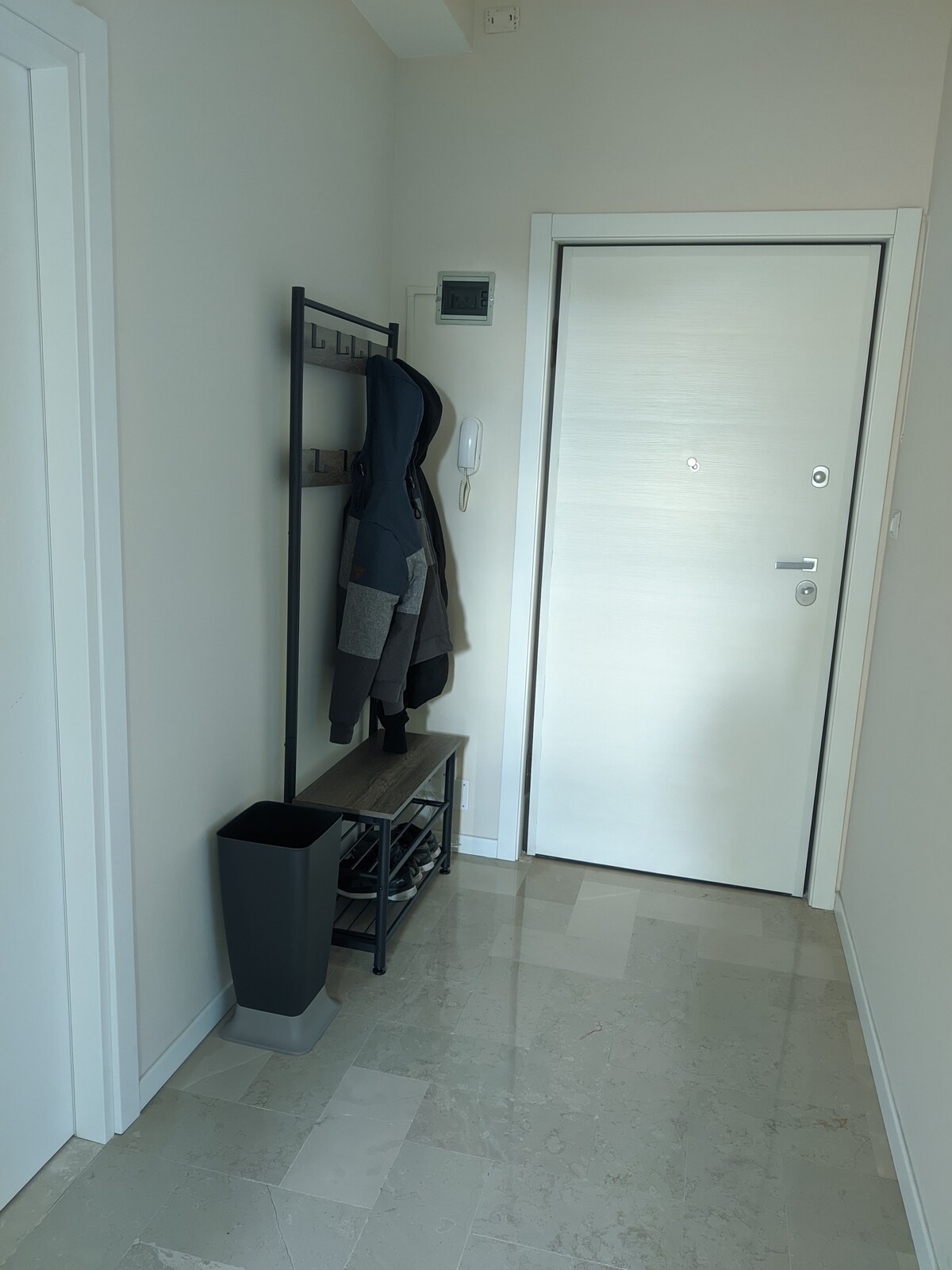 A hallway is presented with light-colored walls and a tiled floor. A coat rack is positioned on one side, holding a jacket. A small shelf unit stands beneath the rack, and a dark container is placed beside it. A secure entrance door is visible.