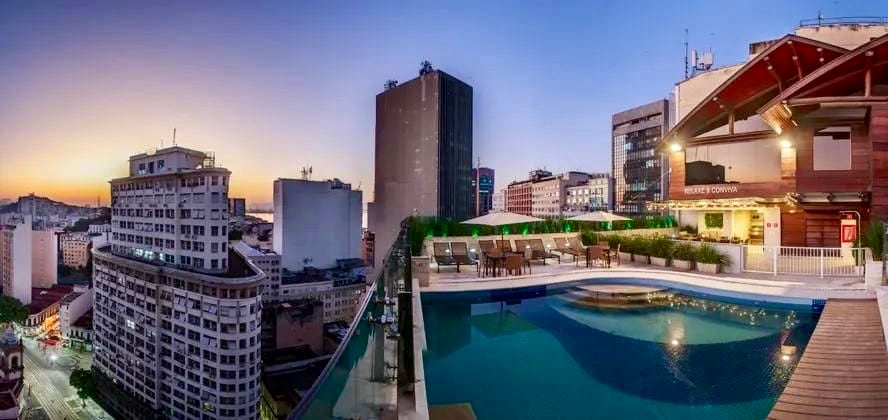 The rooftop pool area is visible, featuring sun loungers arranged along the edge. A wooden deck surrounds the pool, which reflects the soft colors of the sunset. The city skyline, with a mix of modern buildings, is seen in the background.