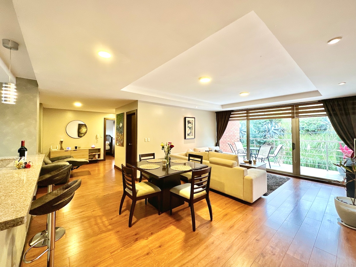 An open living and dining area features a dining table surrounded by chairs, a comfortable sofa, and large windows overlooking greenery. The space is illuminated by soft lighting, with a bar area and high stools positioned nearby, enhancing the inviting ambiance.