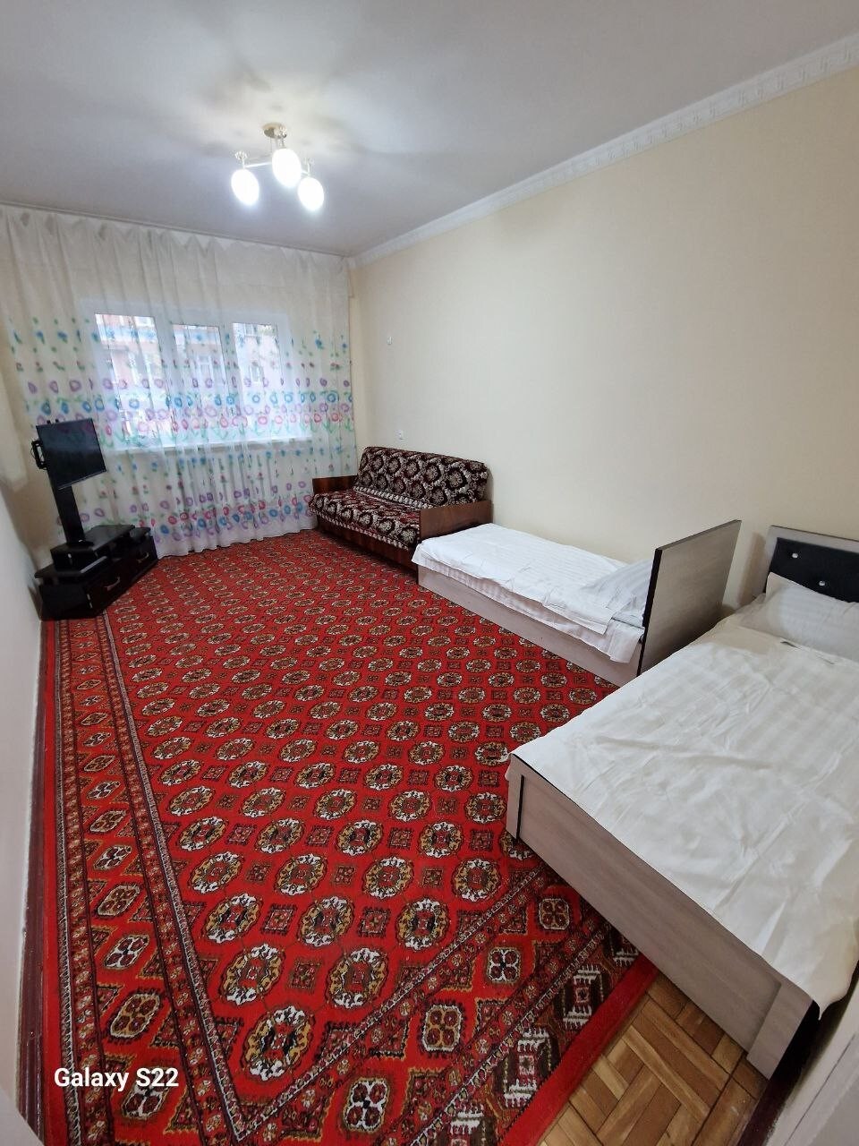 A spacious room is shown with a patterned red carpet covering the floor. A cozy sofa and two single beds are arranged thoughtfully within the space. Large windows are adorned with sheer curtains, allowing natural light to brighten the interior.