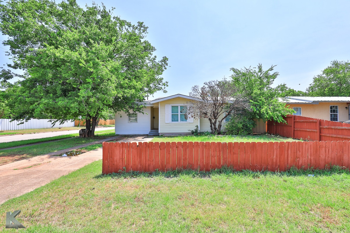 The exterior of a single-story cottage-style home is presented, featuring a well-maintained yard with a vibrant green lawn. A wooden fence encircles the front yard, while trees provide shade. The driveway accommodates parking, and the front entrance is easily accessible from the yard.
