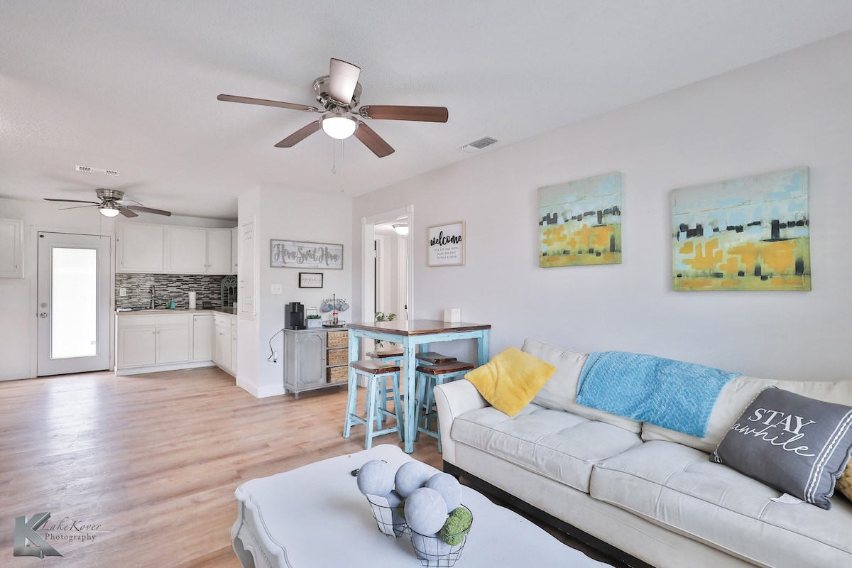 A light and airy living space is highlighted by a cozy white sofa adorned with throw pillows. A small dining area features a blue table and chairs, while a modern kitchen can be seen in the background. Ceiling fans promote airflow, enhancing comfort.