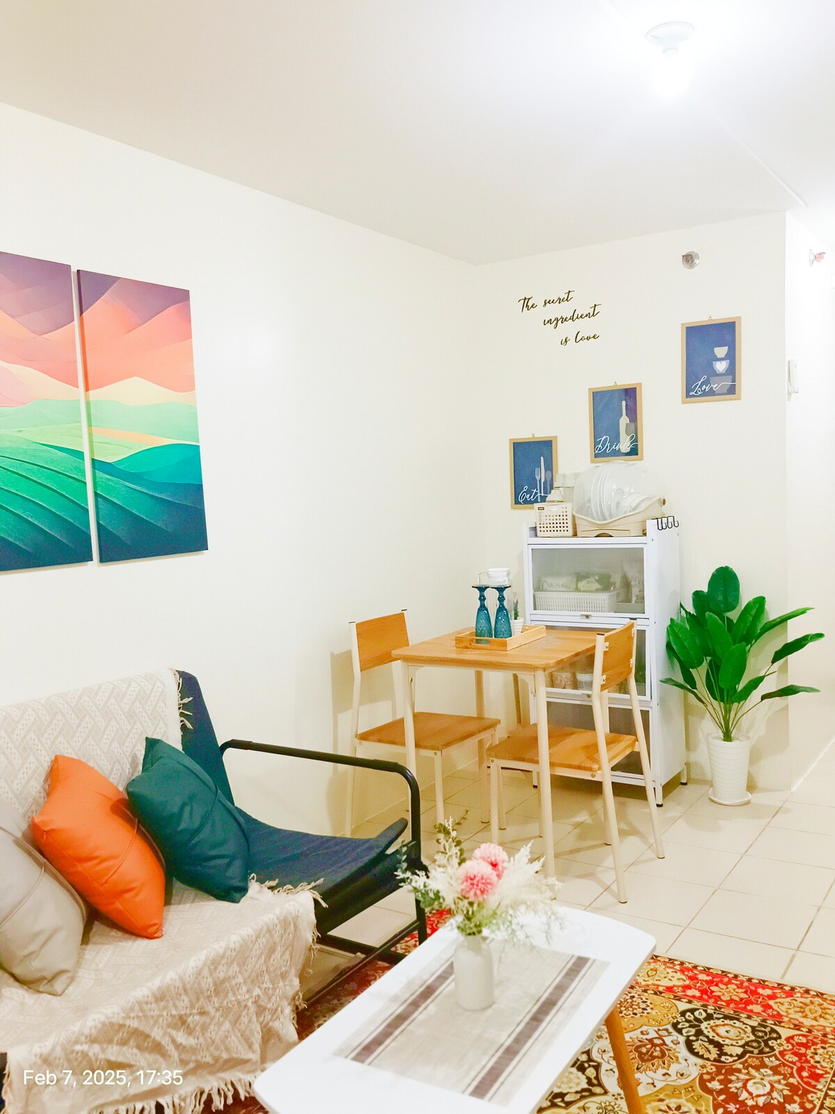 A cozy seating area features a two-seater sofa bed and a small dining table with two chairs. A minimalist white cabinet is positioned against the wall, alongside decorative wall art and potted plants, contributing to a welcoming ambiance.