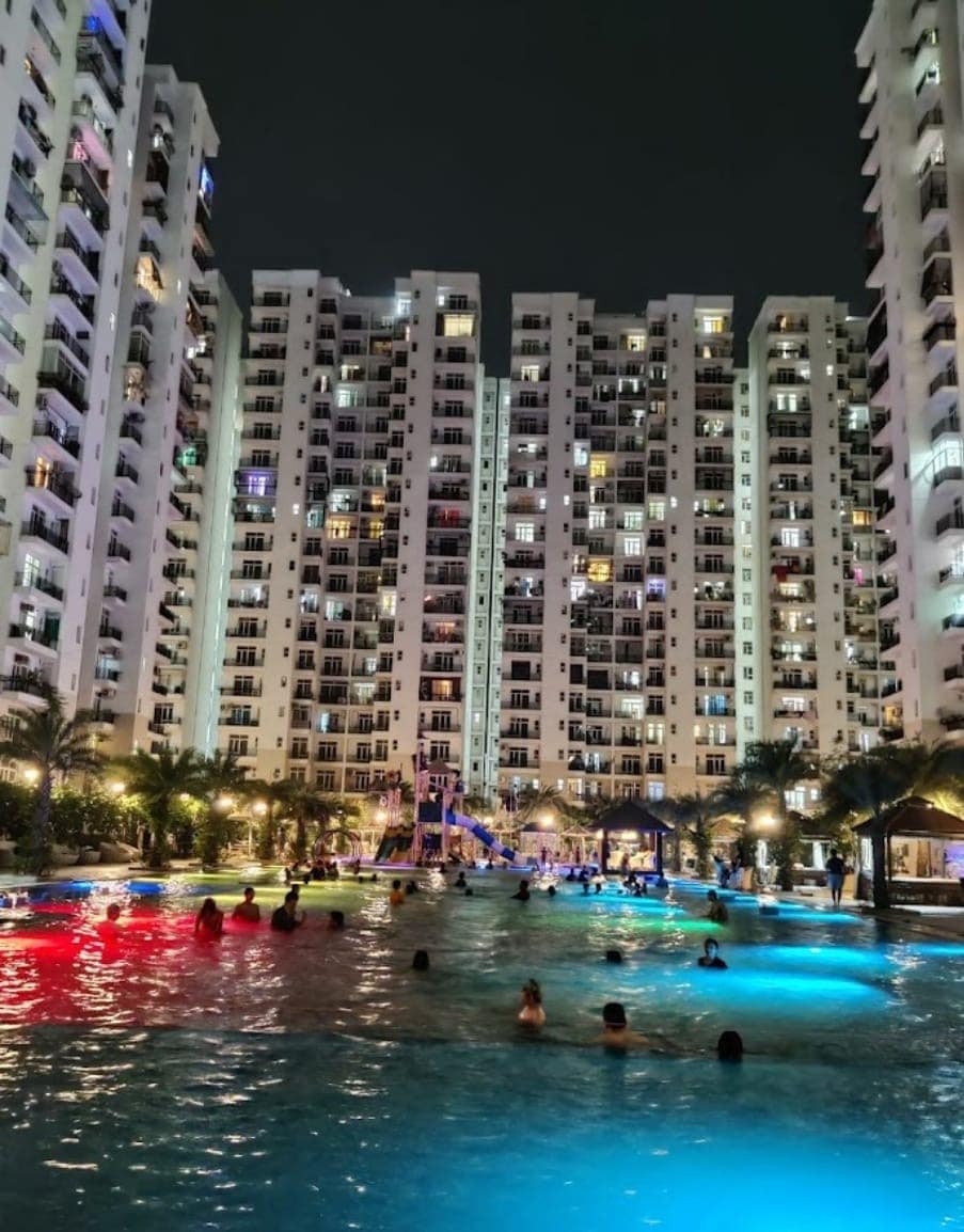 An expansive swimming pool is surrounded by several multi-story residential buildings, with soft lighting creating a relaxing ambiance. Guests can be seen enjoying the water, while colorful lights reflect in the pool. Palm trees add to the tropical atmosphere, enhancing the outdoor experience.