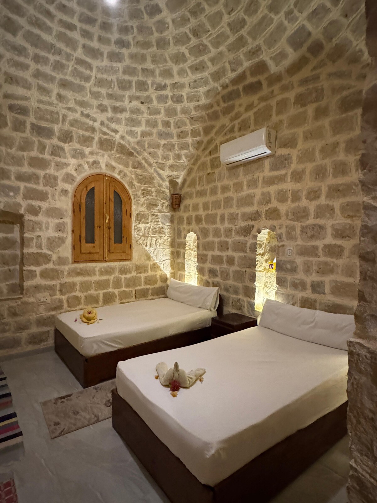 A cozy guest room features two spacious beds covered with light-coloured linens, set against a backdrop of natural stone walls. Wooden window frames and subtle lighting create a serene ambiance. Air conditioning is visible, ensuring comfort in the inviting space.