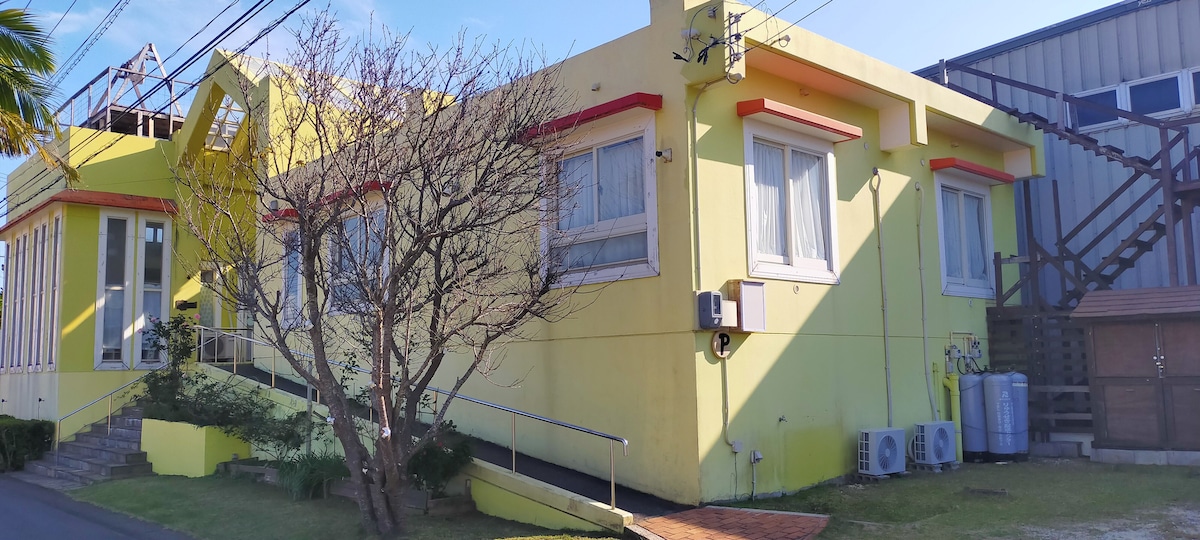 A vibrant yellow building features two prominent wings, highlighting large windows that allow for natural light. Accessibility ramps are visible, supporting ease of entry. Surrounding greenery adds a touch of nature to the setting, complemented by a clear blue sky.