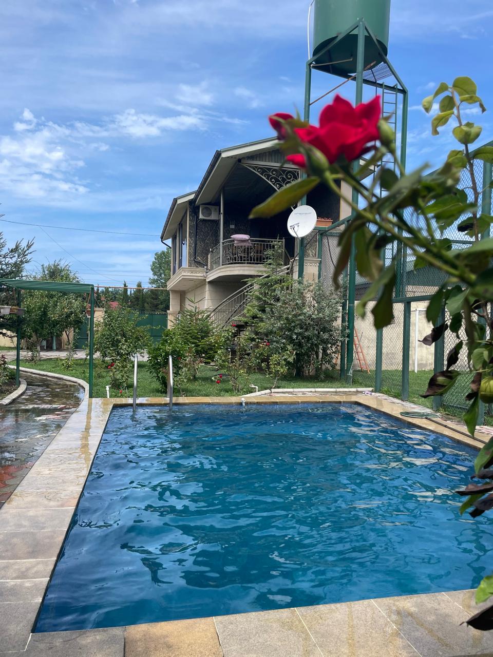 A clear swimming pool is surrounded by stone tiles and manicured greenery, providing a refreshing outdoor space. The villa's two-story structure is visible in the background, featuring a balcony and multiple air conditioning units. A rose bush adds a touch of color in the foreground.
