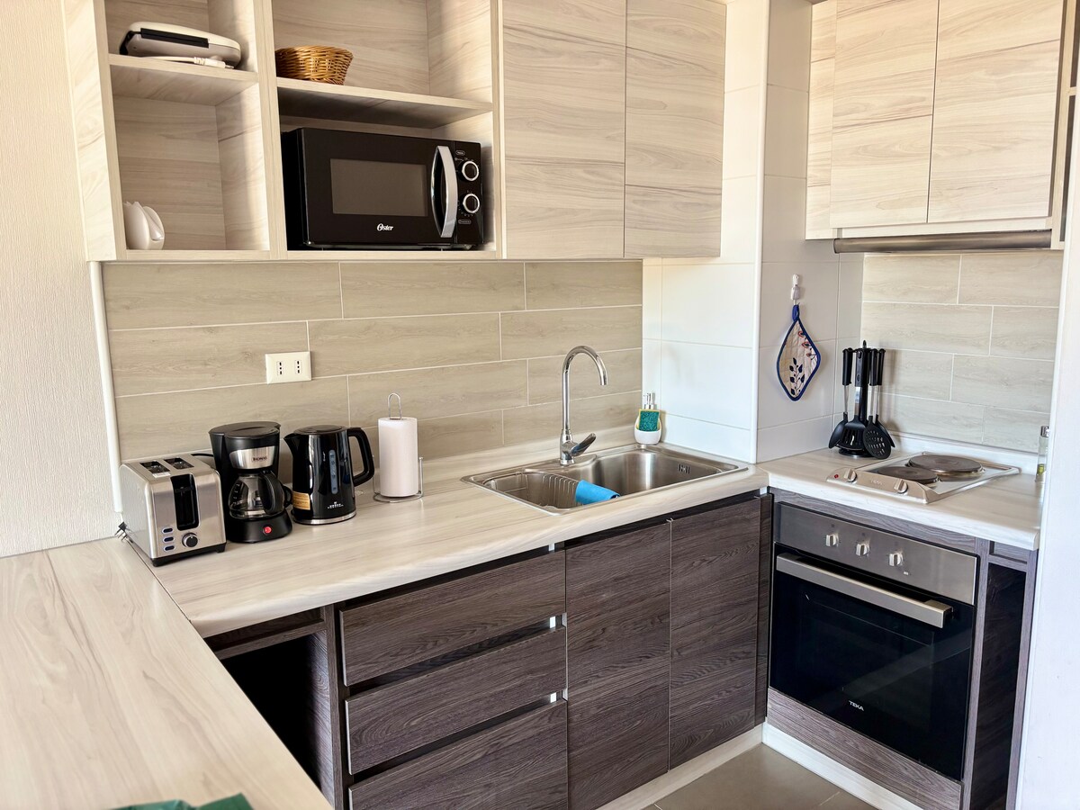 A compact kitchen features modern cabinetry and sleek countertops. Essential appliances, including a coffee maker, kettle, and microwave, are visible. A stainless-steel sink is centrally located, accompanied by cooking utensils. Natural light enhances the overall functionality of the space.