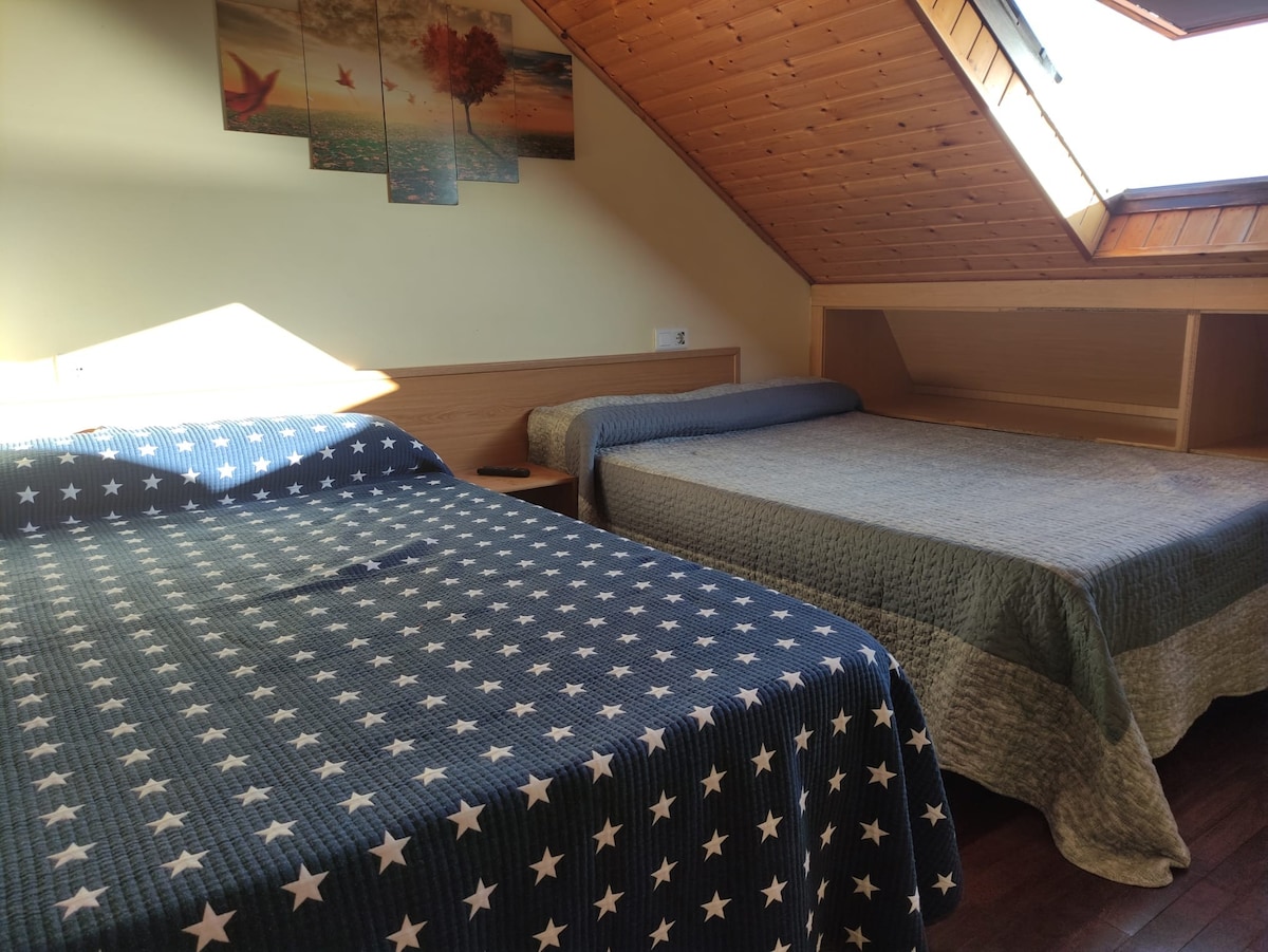 A cozy bedroom is presented with two beds adorned with star-patterned and solid-colored quilts. Natural light filters in through an angled window, highlighting wooden paneling on the ceiling. A small bedside table and a landscape painting add to the inviting ambiance.