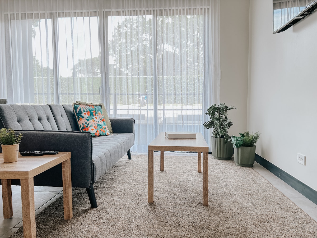 A cozy living area features a gray tufted sofa adorned with colorful throw pillows. A light wood coffee table occupies the center, placed on a soft, beige rug. Potted plants are arranged beside large glass doors, which allow natural light to fill the space.
