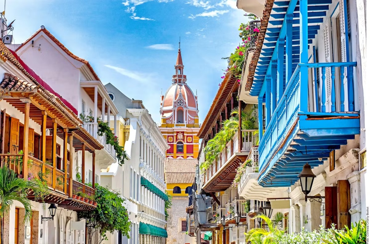 A charming street scene in Cartagena features colorful colonial buildings with blue and white accents. Lush greenery adds a touch of nature, while the iconic clock tower is visible in the background, creating a sense of the city's rich architectural history.