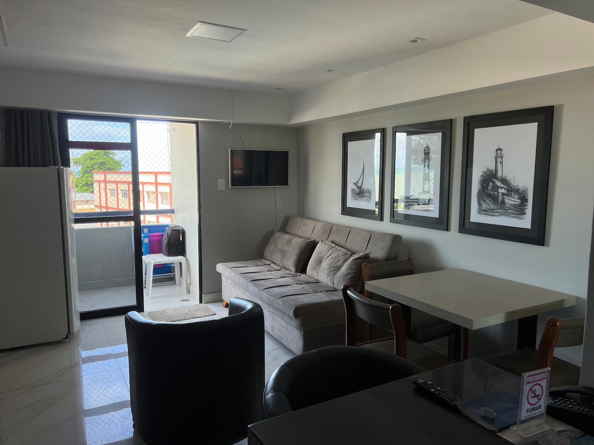 A comfortable living area is observed, featuring a spacious sofa and a dining table with chairs. Natural light enters through a balcony door, complemented by framed artworks on the wall. A flat-screen television is mounted nearby, providing convenient entertainment options.