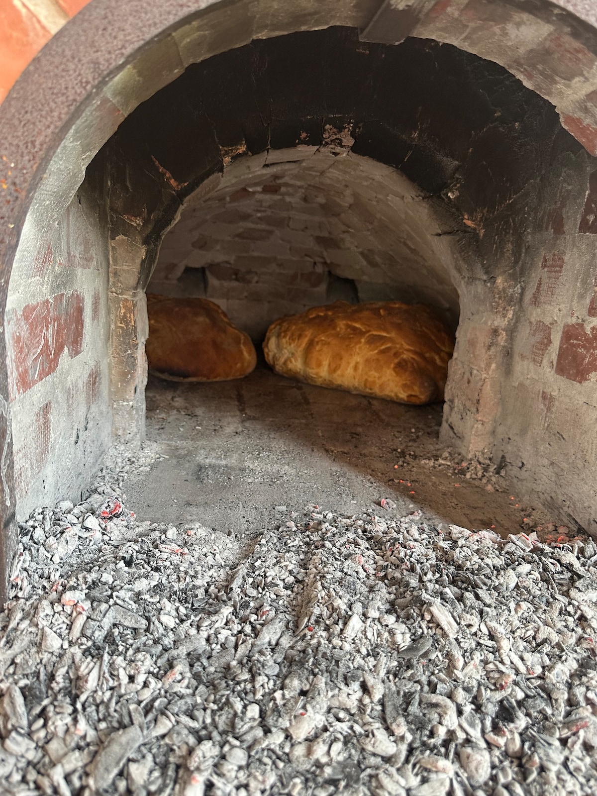 Two golden-brown loaves of bread are nestled inside a traditional brick oven. The textured walls of the oven are visible, framed by a crescent opening. Charred ash and remnants of fire cover the floor, suggesting recent use.