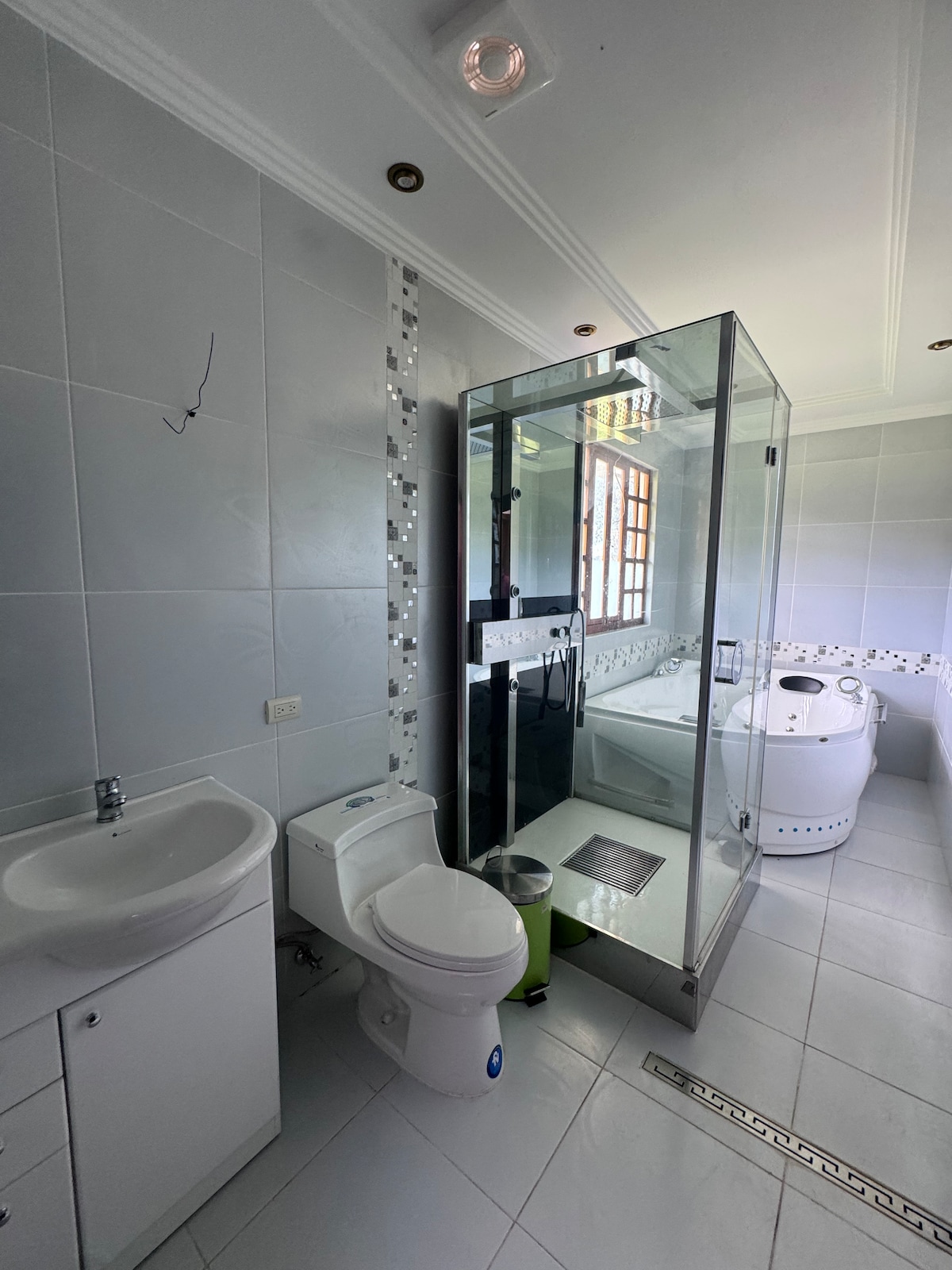 A modern bathroom is displayed, featuring a glass shower enclosure alongside a bathtub. White tiles cover the walls and floor, creating a clean and bright space. A small vanity with a sink is visible, complemented by a mirror above. Natural light enters through a nearby window.