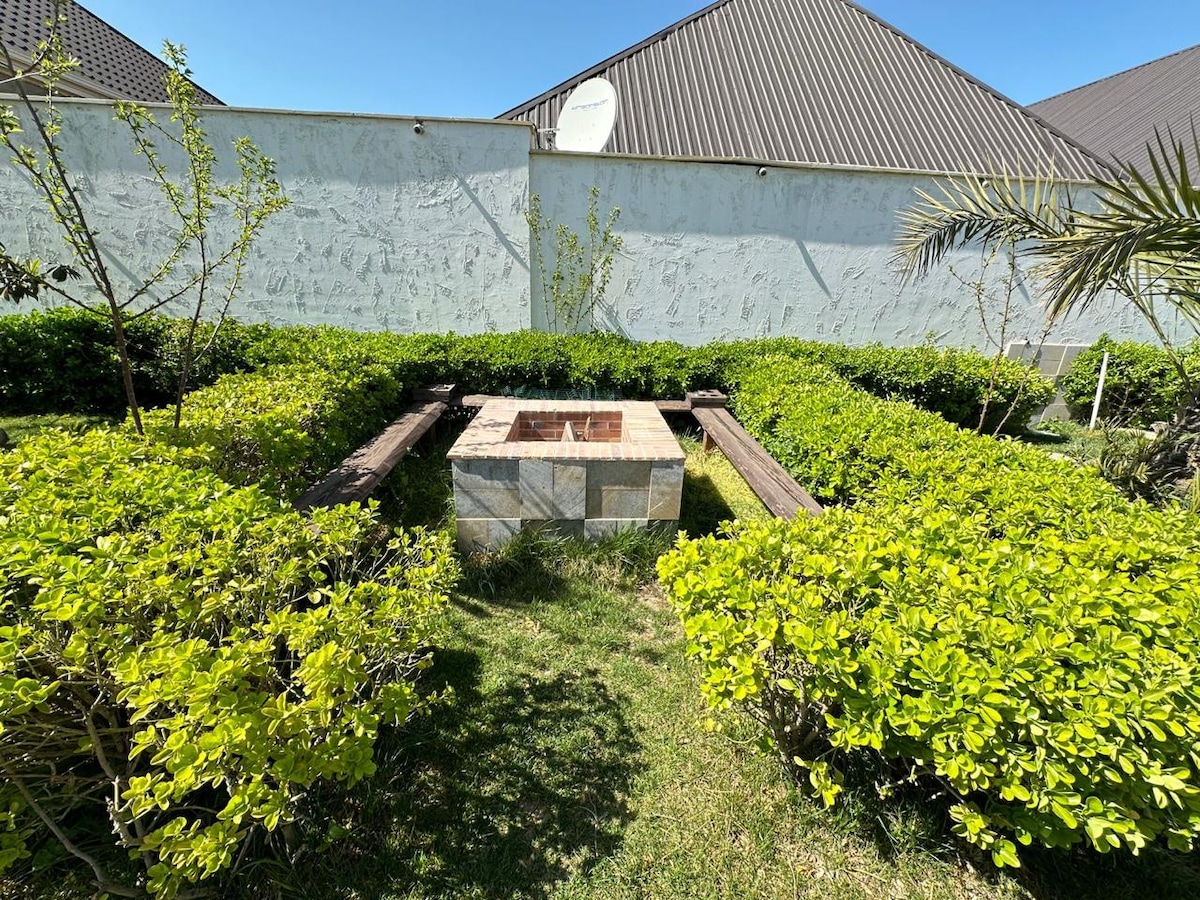A designated outdoor fire pit is situated within a landscaped area, surrounded by vibrant green bushes and low shrubs. Wooden benches are placed around the stone structure, providing a communal space for guests to gather. The clear blue sky complements the serene setting.