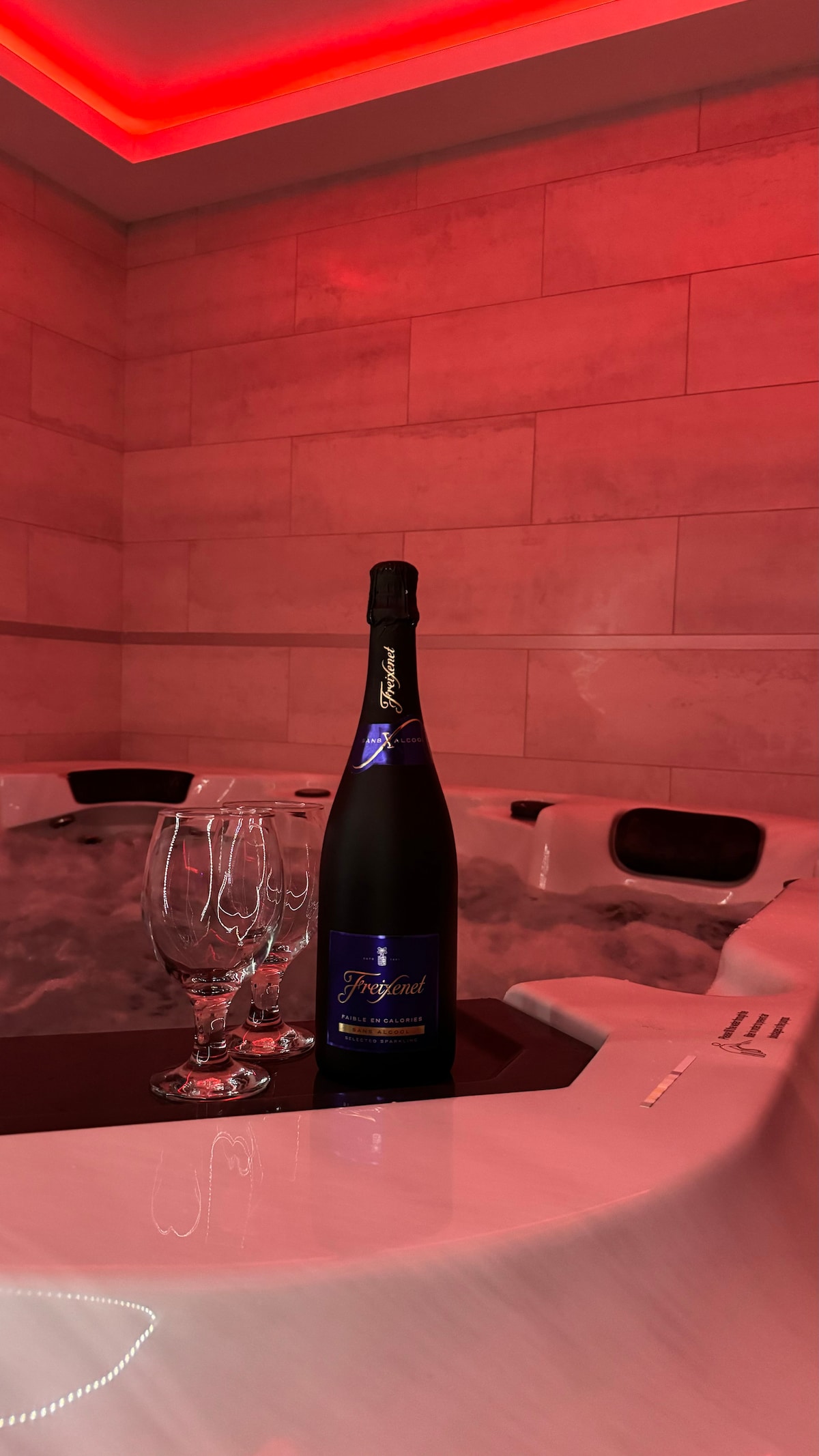 A jacuzzi is shown with soft, ambient red lighting, creating a relaxing atmosphere. A bottle of sparkling wine is positioned beside two elegant glasses, hinting at a serene moment of indulgence. The smooth surface of the jacuzzi adds to the space's modern aesthetic.