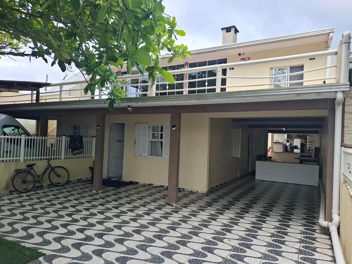 A spacious two-story house is presented, featuring a covered parking area adorned with a patterned tile floor. The upper level is visible, showcasing a balcony with railing. Large windows provide natural light to the interior, while a bicycle is casually placed nearby.
