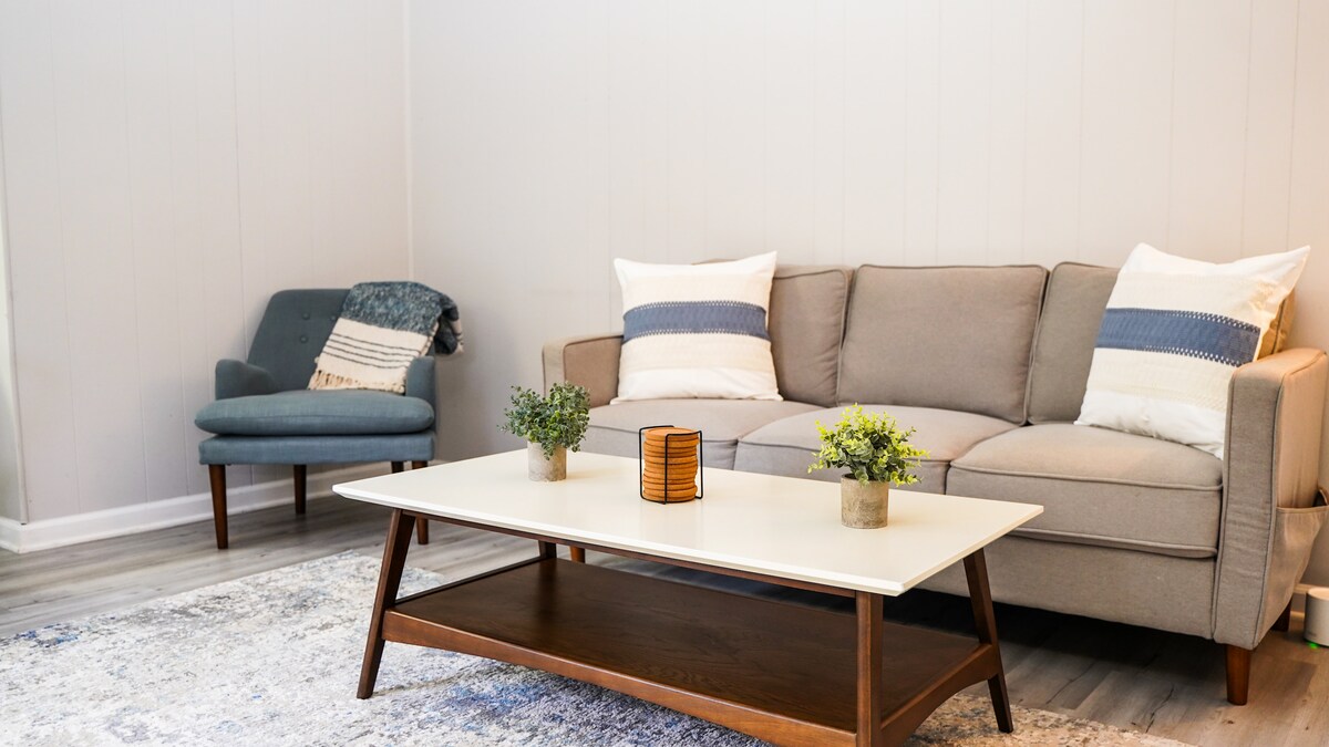 The living room features a comfortable couch adorned with soft pillows, complemented by a cozy chair. A coffee table sits at the center, decorated with small plants. Light-colored walls create a bright and airy atmosphere, enhanced by a patterned area rug underfoot.