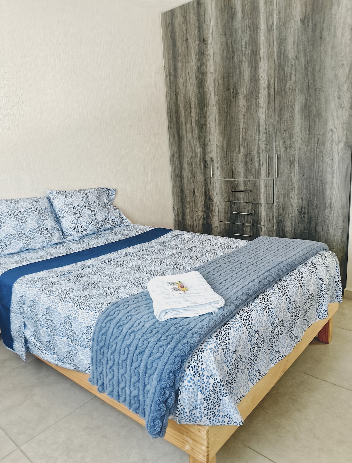 A sleeping area is equipped with a spacious bed dressed in a patterned blue and white coverlet. A textured blue throw and neatly folded towels are placed at the foot of the bed. A tall wardrobe with wood-like finish is positioned against the wall.