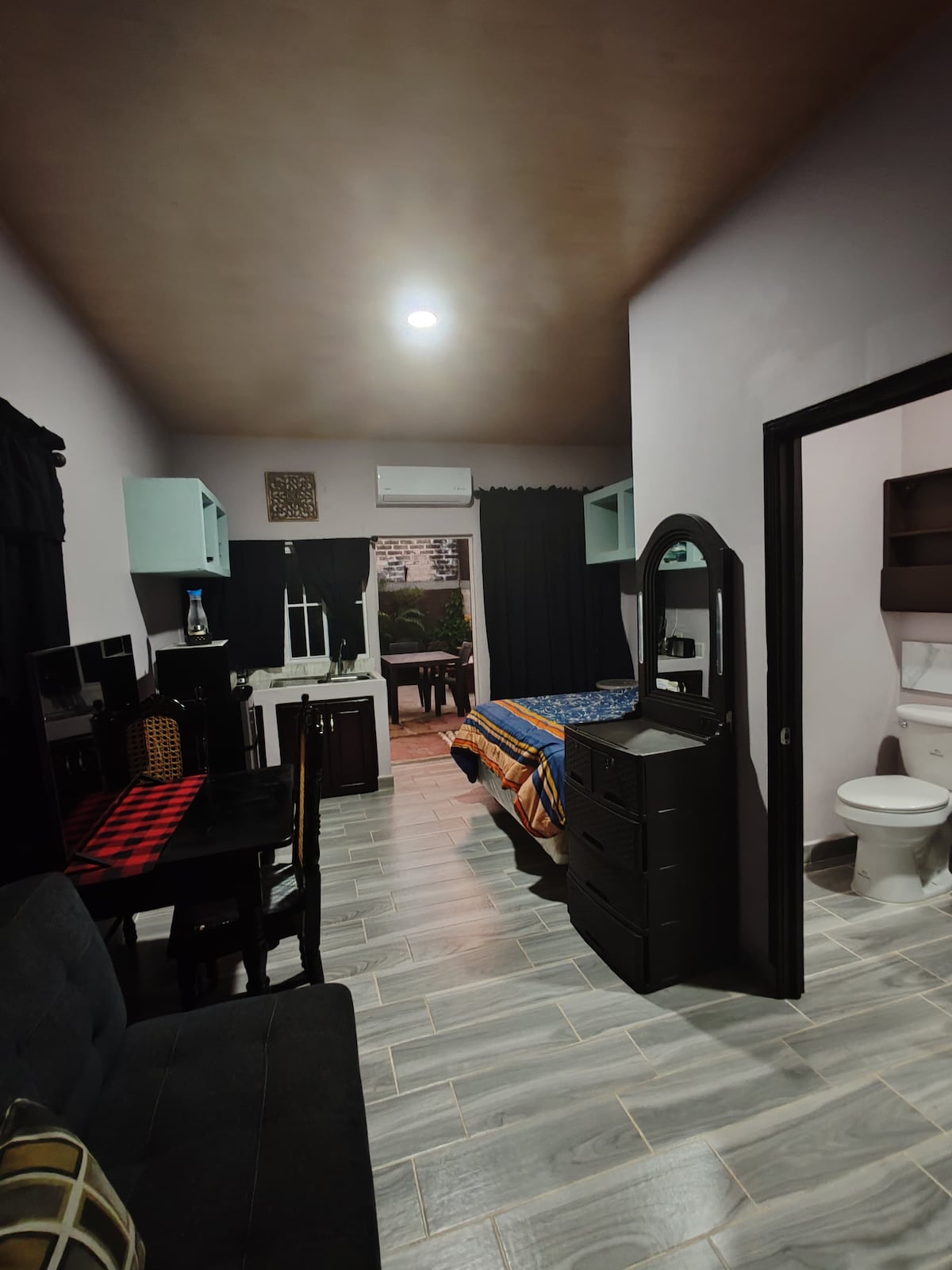 A private guest suite features a spacious bed with colorful bedding, paired with a sleek dresser and a small dining area. A comfortable sofa sits nearby. The bathroom is visible with modern fixtures. Natural light enhances the cozy atmosphere.
