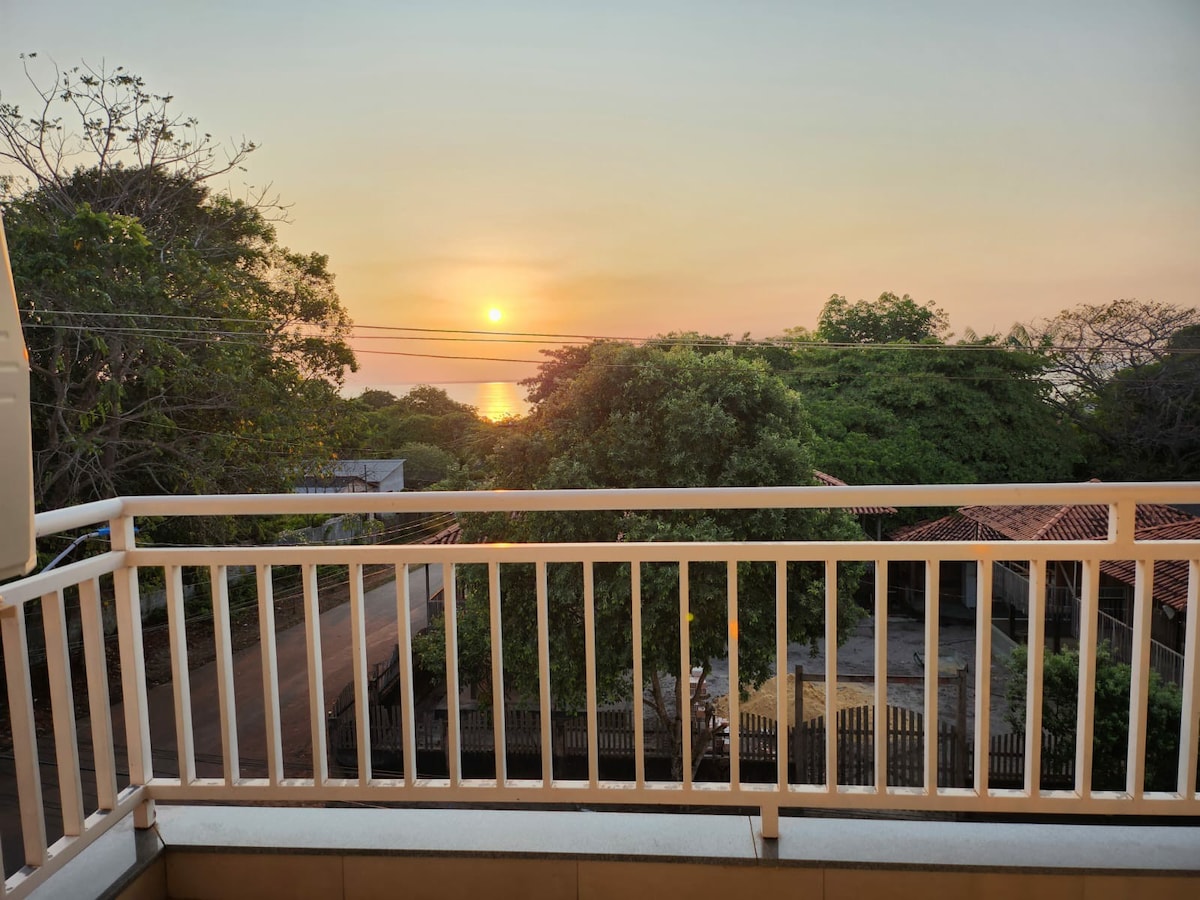 The image captures a sunset view from a balcony, where the sun sets over the water, casting warm hues across the sky. Lush trees frame the scene, and rooftops of nearby buildings are visible in the foreground, highlighting a tranquil ambiance.