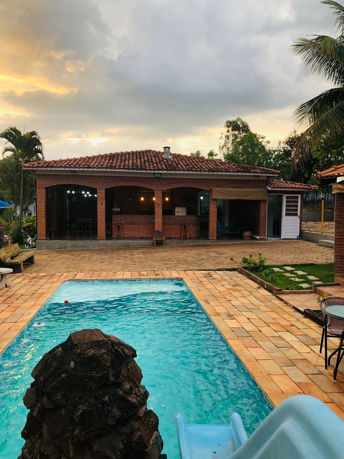 The image displays a charming outdoor space featuring a swimming pool with a rock fountain. A spacious patio with a brick house is visible in the background, accompanied by garden areas and palm trees. The sky presents a soft, golden hue as the sun sets.