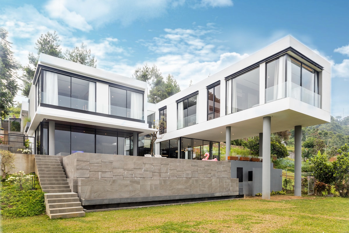 A modern villa is showcased with a striking architectural design, featuring multiple levels and expansive glass windows. The exterior includes a stone terrace and steps leading down to the well-maintained lawn, surrounded by lush greenery and scenic hills.