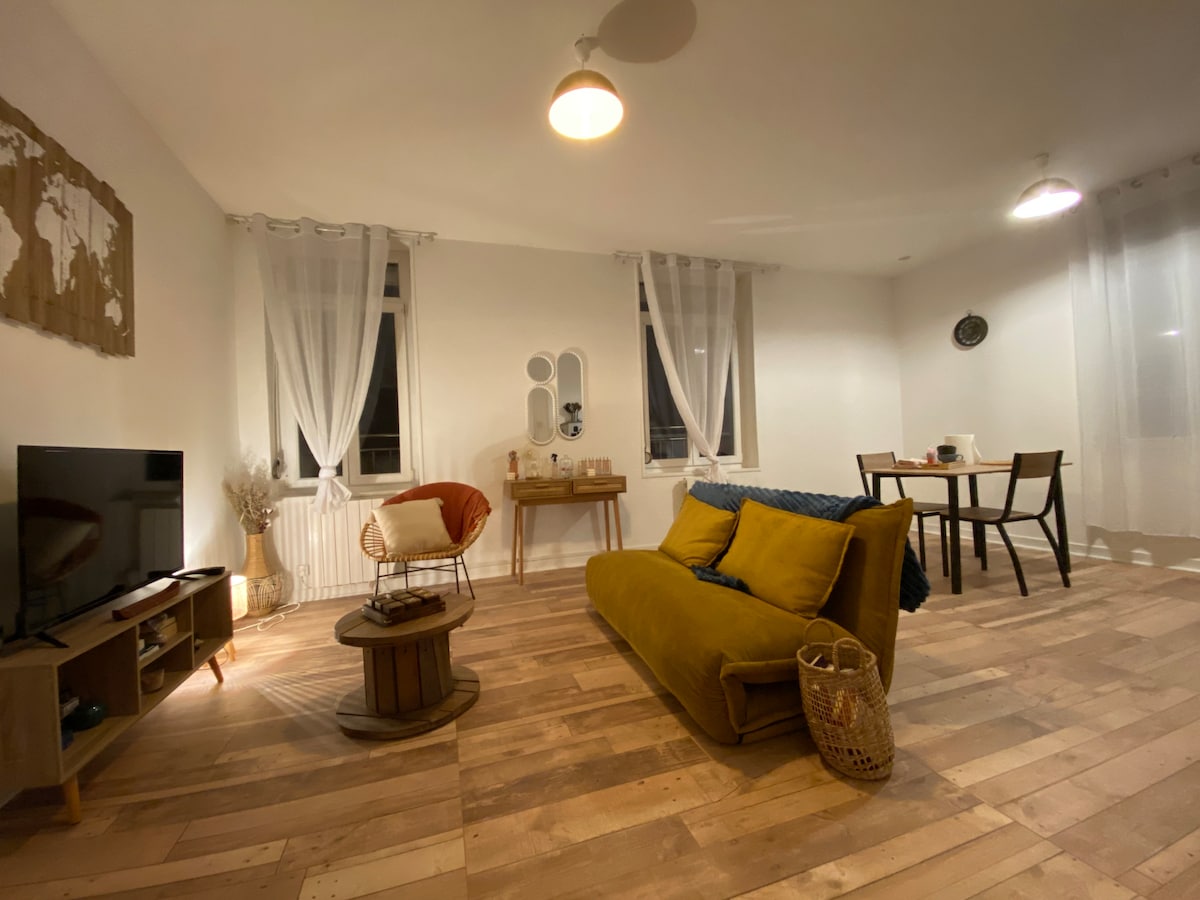 The living area features a plush sofa and a round table with four chairs. A television is positioned on a wooden stand, while large windows with sheer curtains provide natural light. Decorative elements include a globe and a woven basket, enhancing the space's simplicity.
