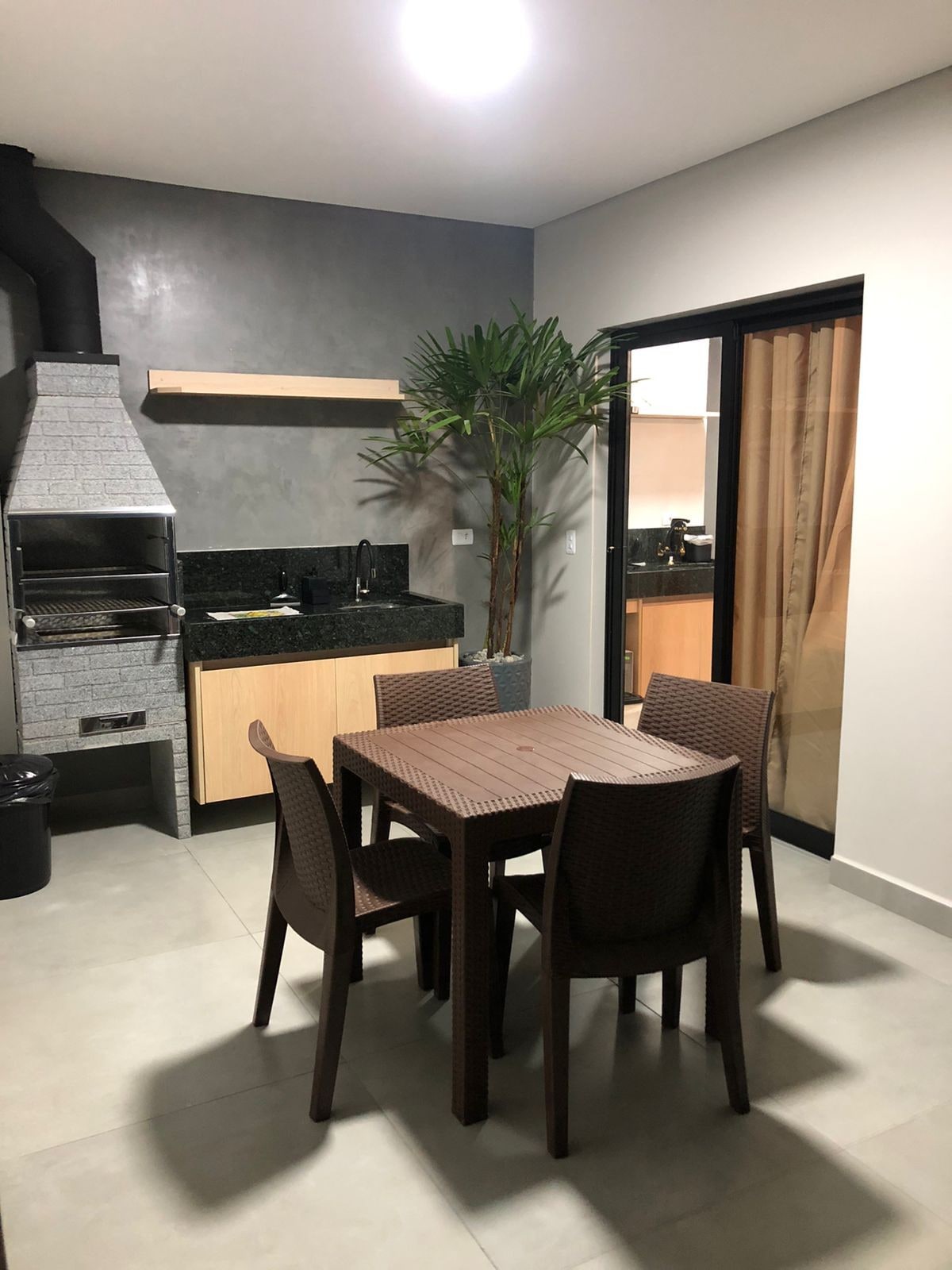 A dining area is featured with a wooden table and four matching chairs, accompanied by a built-in grill and a compact kitchen counter. A potted palm adds a touch of greenery, while large glass doors provide natural light and access to the outdoor space.