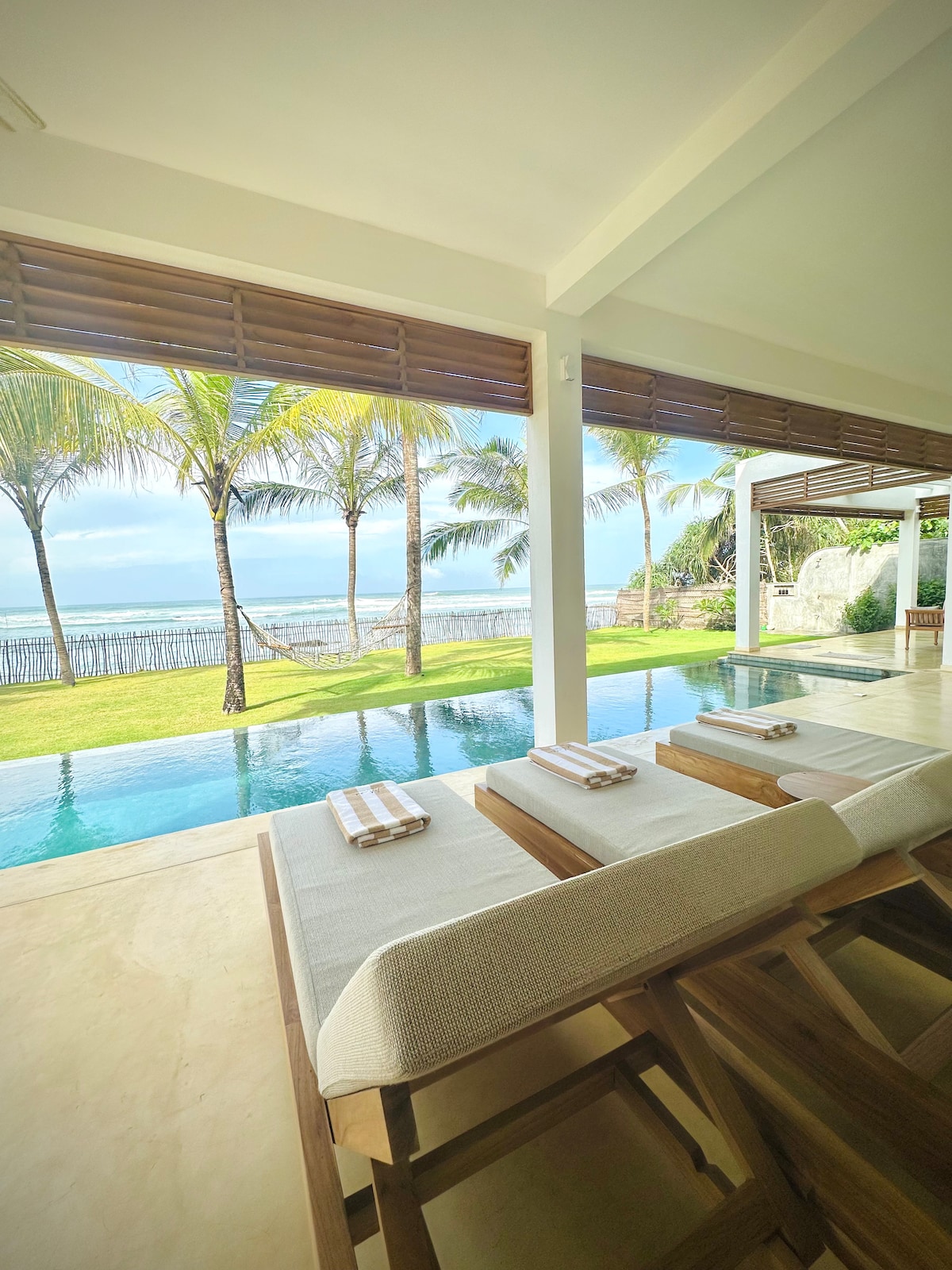 An open living space overlooks a serene infinity pool, framed by lush palm trees. Natural light floods the area, with a view of the ocean visible through spacious glass doors. Comfortable seating is arranged thoughtfully for relaxation and enjoyment of the coastal scenery.