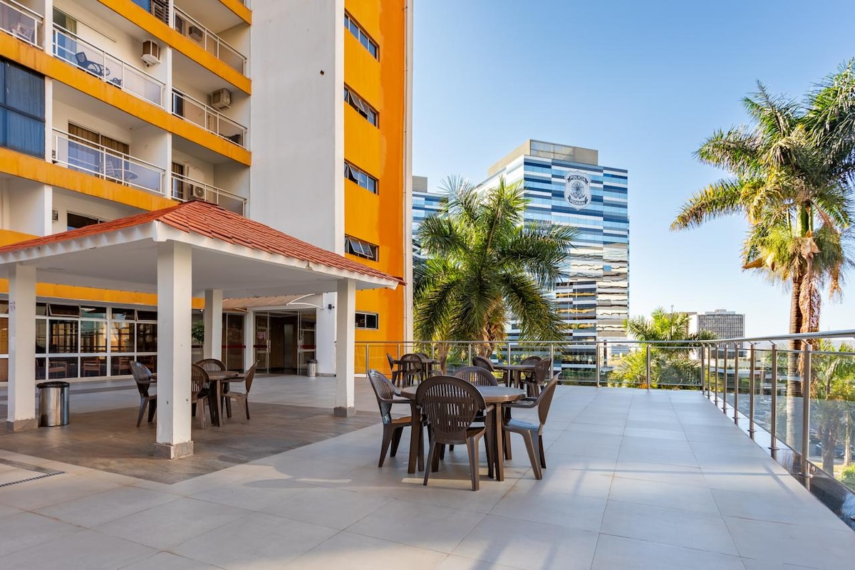 An outdoor terrace is presented with several round tables and chairs, surrounded by palm trees and modern buildings. The area is designed for relaxation, with a covered gazebo offering shade, while clear skies provide a bright ambiance.