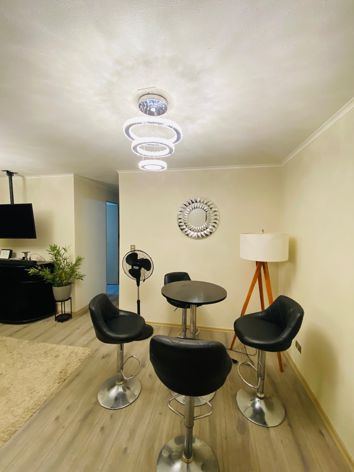 A modern dining area features a round table surrounded by four black bar stools with metallic bases. A stylish light fixture hangs from the ceiling, providing soft illumination. A decorative mirror reflects light on one wall, complemented by a small plant and a floor lamp nearby.