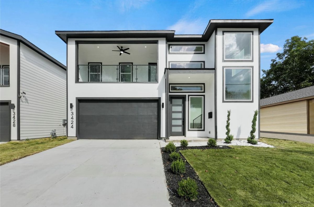 A modern two-story house features a sleek facade with large windows and a minimalistic design. A spacious driveway leads to a dark garage door, and neat landscaping with small shrubs frames the entrance. A covered balcony with glass railings is visible on the second floor.