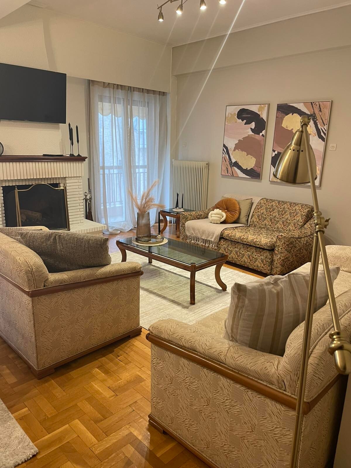 The living area features a light-colored sofa set arranged around a coffee table. Two armchairs complement the seating arrangement. Large windows allow natural light to fill the space, while two pieces of wall art enhance the decor. A floor lamp stands nearby, providing additional lighting.