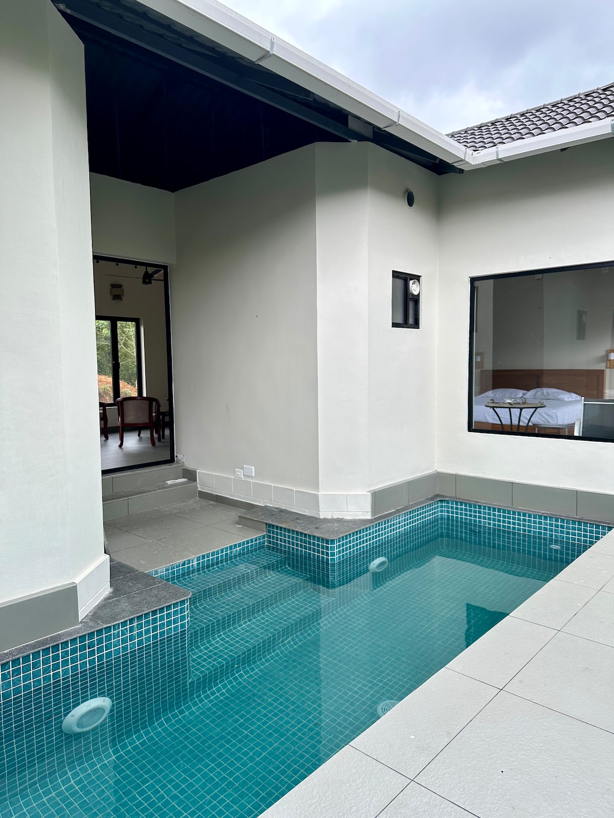 A private pool is situated within a minimalist design, featuring clear blue water and sleek tiles. The pool area connects to an open space, which leads to a well-lit living area visible through an expansive window, enhancing the sense of spaciousness.