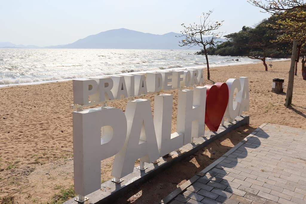 A coastal sign reading 'Praia de Fora Palhoça' stands on a sandy beach. The background features gentle waves lapping against the shore, with distant mountains providing a scenic backdrop. Sparse trees dot the area, offering a hint of shade along the pathways.