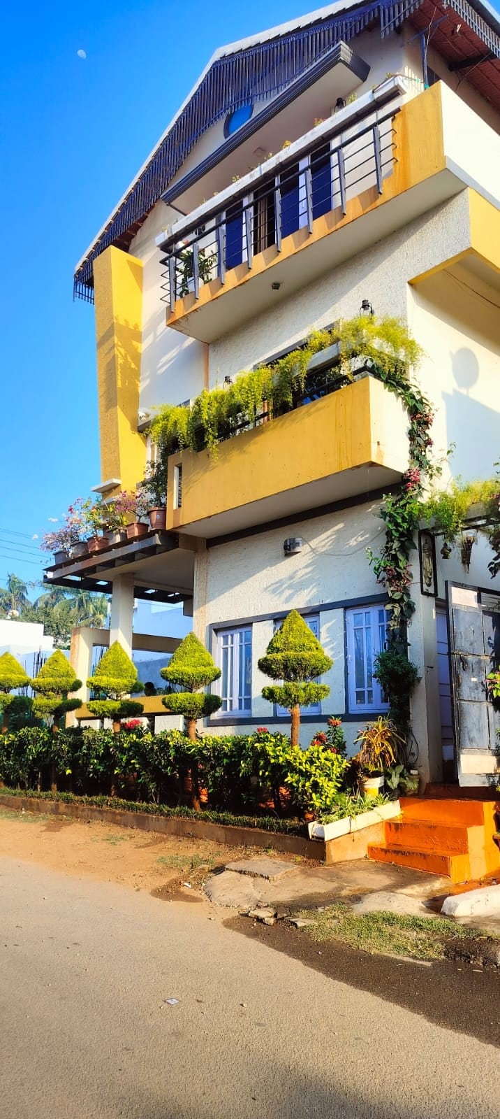 A three-story building features a bright yellow exterior complemented by decorative greenery. Floral arrangements and neatly manicured shrubs provide a welcoming entrance. Large windows allow natural light, while a pathway leads to the main entrance, framed by lush vegetation.