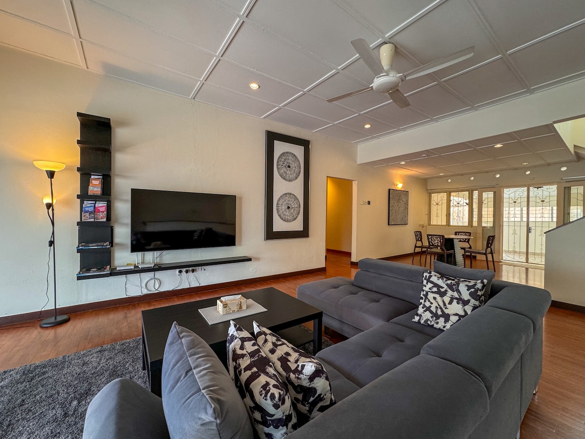 The living room features a large sectional sofa adorned with patterned pillows, positioned in front of a 65-inch Samsung smart TV mounted on the wall. A sleek coffee table is centered in the space, complemented by a floor lamp, and ample natural light fills the room through several windows.