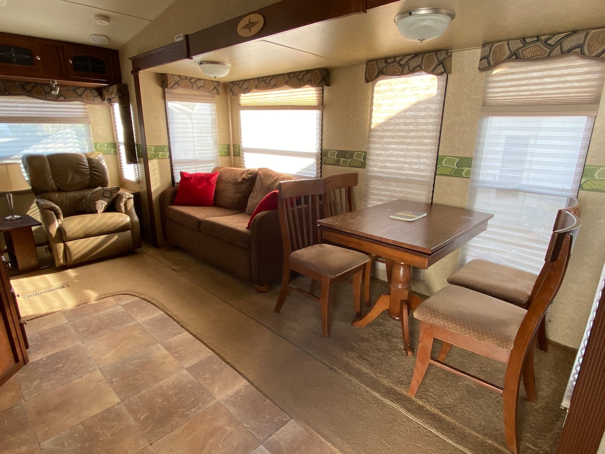 A compact living space is featured, with a comfortable couch adorned with a red accent pillow. A small dining table is accompanied by two wooden chairs, while a recliner is positioned nearby. Natural light filters through the window coverings, illuminating the area.
